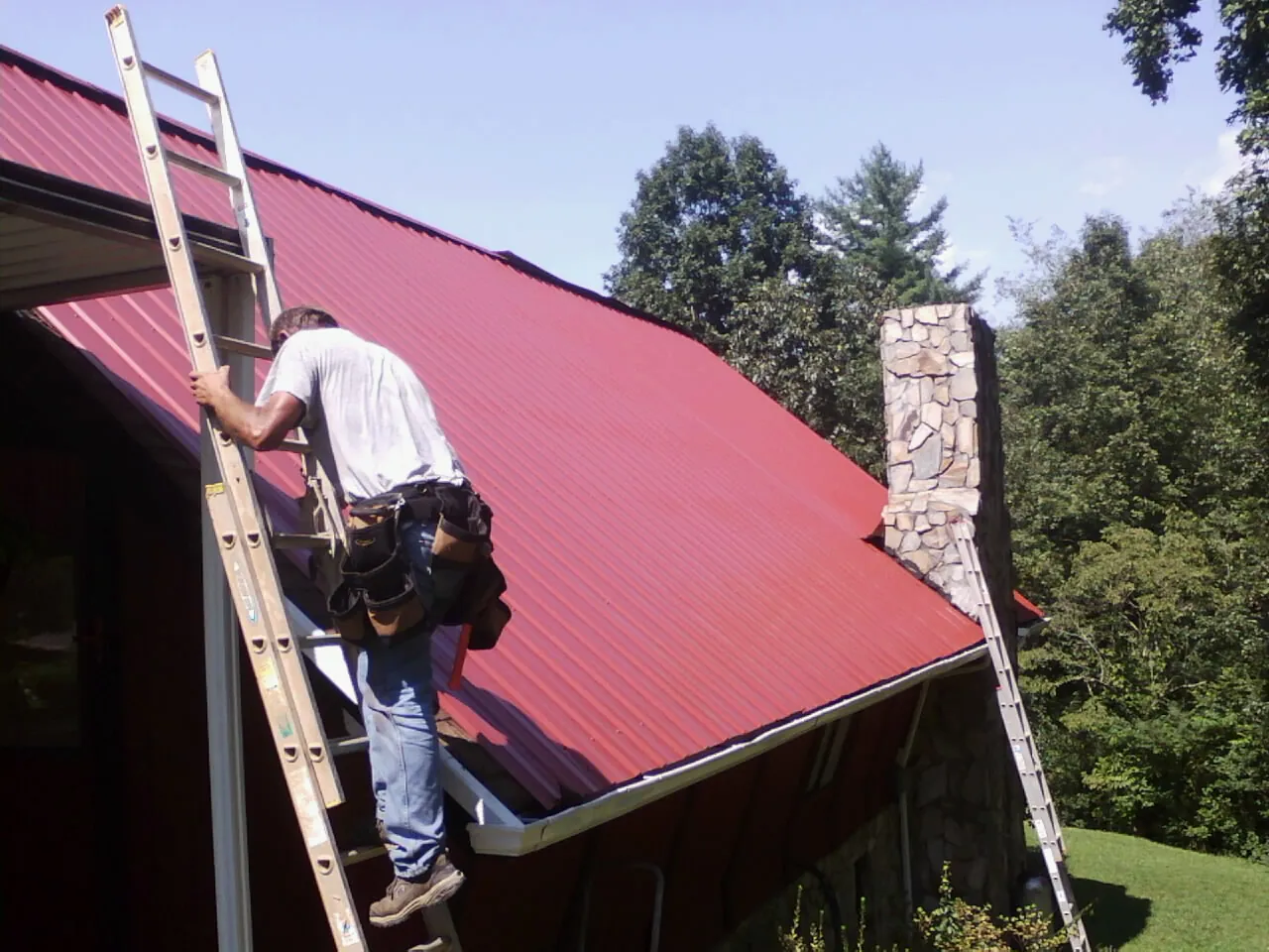 Roofer on a ladder beside a red metal roof and stone chimney — Soffit & Fascia Repair by Roofing Brothers in Street